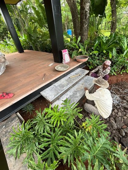 Workers lay heavy stone slabs to create steps leading to a wooden deck. This process of integrating hardscaping with the surrounding lush greenery is key to creating a harmonious outdoor space.