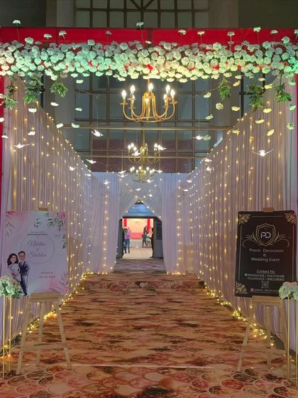 A classic and elegant entrance gate design. We used a bold red frame, a canopy of white flowers, and a walkway lit with fairy lights and chandeliers to create a warm and welcoming path for guests.