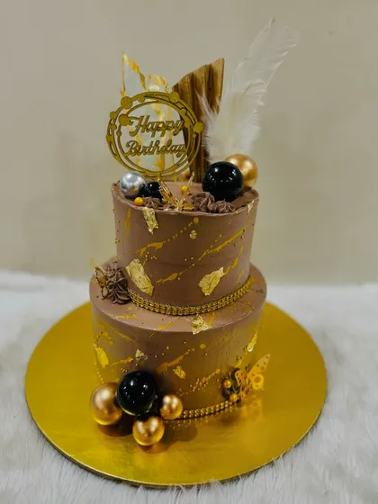 A sophisticated two-tiered chocolate cake perfect for an adult celebration. The combination of rich chocolate, gold leaf, and a modern feather topper creates a look of pure luxury.