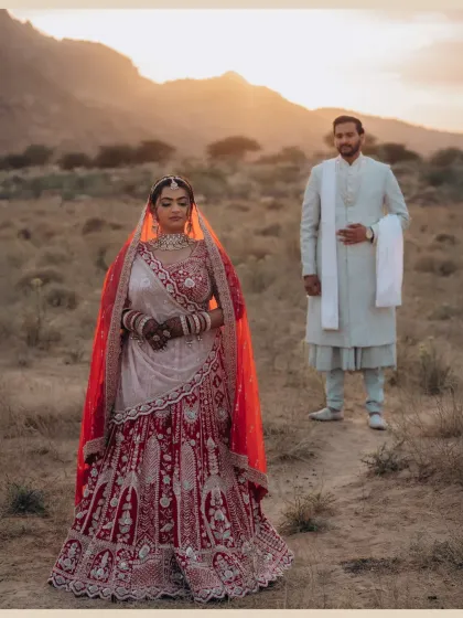A stunning portrait of Nishi against the warm, setting sun. The golden light beautifully highlights the intricate details of her red lehenga, set against the rugged mountain landscape.