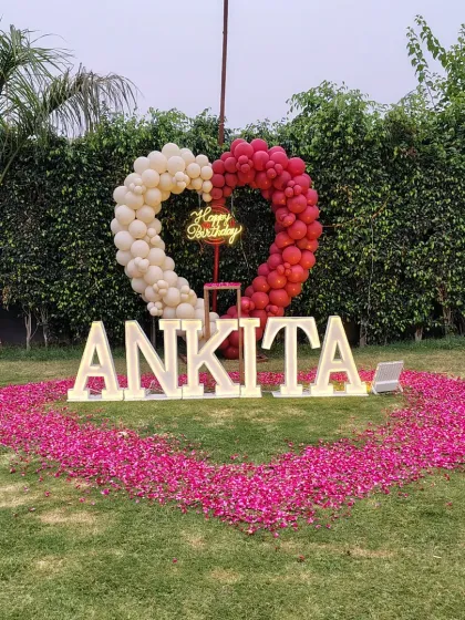 Another look at a grand outdoor birthday celebration for Ankita. This setup shows a heart-shaped balloon arch in red and white, a neon sign, and a heart on the grass made of fresh rose petals, perfect for a big surprise.