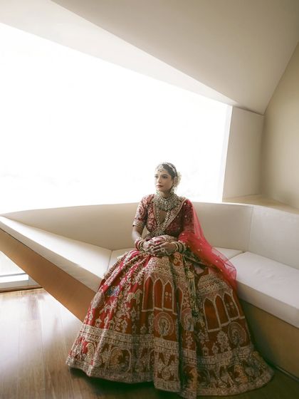 A full-length portrait of the bride seated, showcasing the magnificent embroidery and spread of her red wedding lehenga.