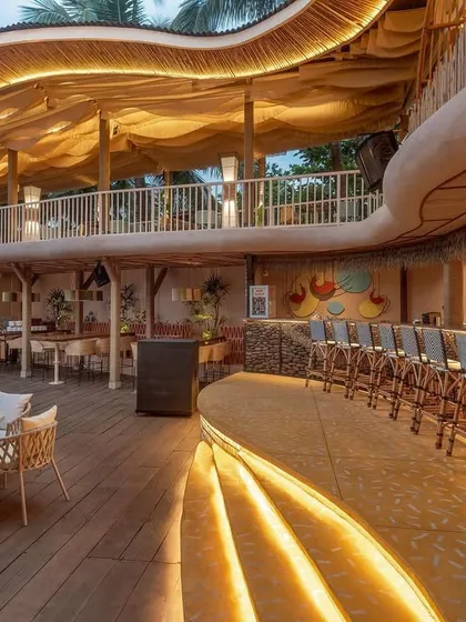 The main bar at Dubki at dusk, with warm lighting highlighting its curved form. The bar is integrated into the lower level, with a bamboo-clad backdrop and woven bar stools that reinforce the project's earthy, sustainable material palette.