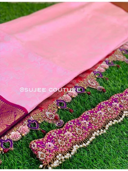 A beautiful flat-lay showcasing the components of a custom half saree. It includes a baby pink saree, a heavily embroidered purple vaddanam, and matching tassels with heart-shaped details.