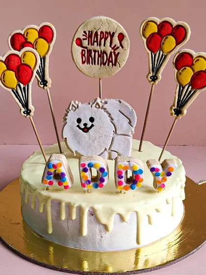 Addy the Pomeranian's cake features a white chocolate drip and a fluffy pom cookie topper. The colorful letter cookies add a touch of fun.