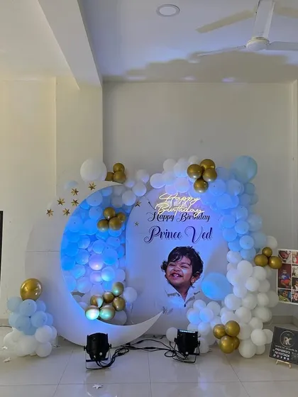 This moon-themed birthday decoration includes a personalized backdrop with the child's photo. The setup is complemented by a milestone photo board, flower vases, and a blue and white balloon garland with gold accents.