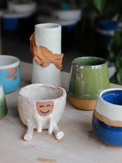 A whimsical collection of student work, featuring a smiling planter on legs, a vase with a sculpted fish, and colorful mugs.