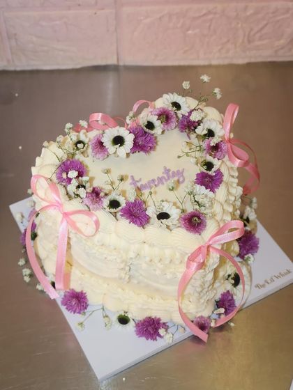 A heart-shaped cake for a garden fairy vibe. This design is covered in intricate piping and adorned with fresh purple and white flowers and delicate pink ribbons.