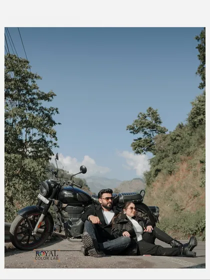 A wide-angle shot of a couple sitting with their motorcycle against a clear blue sky and lush green hills, capturing the freedom of the open road.