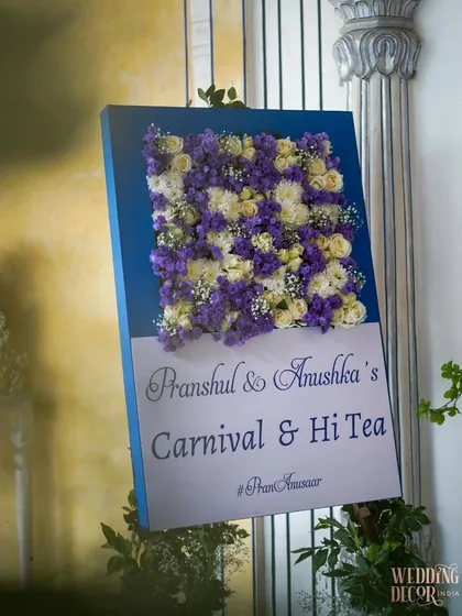 A custom welcome sign for the "Carnival & Hi Tea" event, decorated with a beautiful arrangement of purple and white flowers.