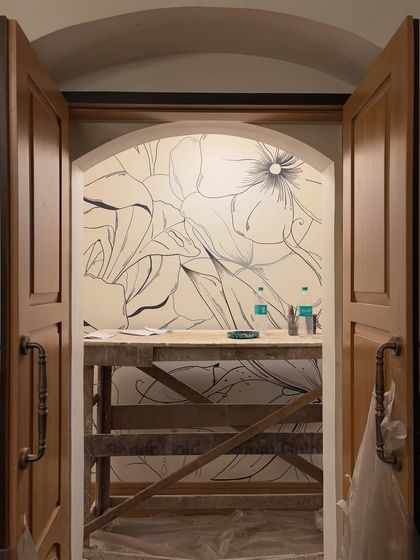 A view of the floral line art mural in progress, seen through a doorway. The scaffolding and unfinished state reveal the reality of creating art on a large scale.