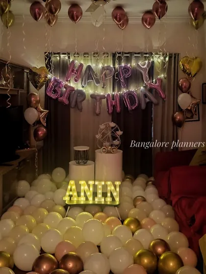 A living room transformed for a birthday surprise, with the floor covered in white and gold balloons, heart-shaped foil balloons, and personalized marquee letters.