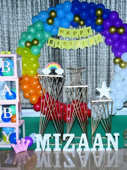 A rainbow-themed birthday party featuring a multi-colored balloon arch and 'BABY' display boxes. The setup uses unique stands for the cake and props.