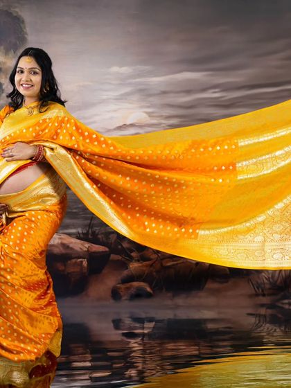 A creative and artistic portrait featuring a mom-to-be in a flowing yellow saree. The painted backdrop and the tossed fabric create a dreamy, ethereal effect.
