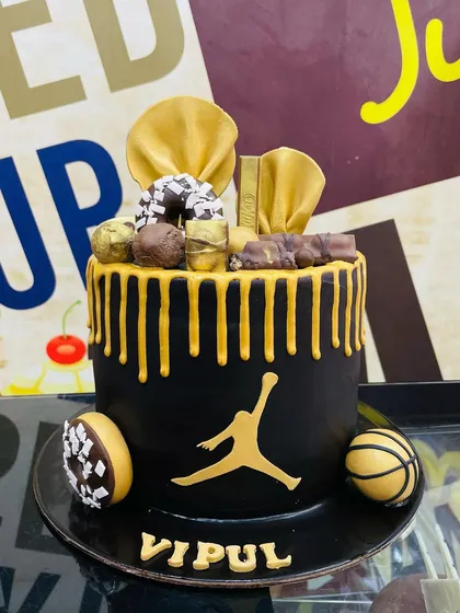 We can design a cake around any theme, like this custom basketball-inspired creation. It features a sleek black fondant finish, a striking gold drip, and is topped with miniature donuts, chocolate bars, and edible basketball details, all centered around the iconic Jordan logo.