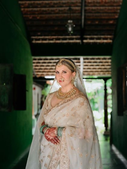 A bride in a white saree, her face framed by her veil, stands in the green corridor, a picture of grace and elegance.