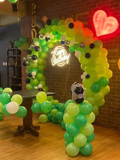 A football-themed balloon arch in a restaurant with a brick wall interior. The green balloons and neon heart sign create a fun and unique look.