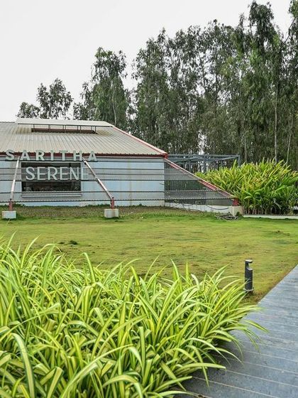 The Saritha Serene marketing office is set within a wide expanse of green lawn. The building's industrial-inspired architecture, with its metal cladding and truss system, contrasts with the soft, naturalistic landscape.