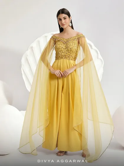 The vibrant Emma Jumpsuit in a brilliant yellow. This piece is designed for graceful movement, featuring an embellished off-shoulder bodice with dramatic cape sleeves and wide-leg pleated trousers for a truly modern festive outfit.