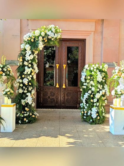 An outdoor wedding entrance featuring a beautiful arch of white flowers and greenery. The design is fresh, natural, and elegant, perfectly complementing the venue's architecture.