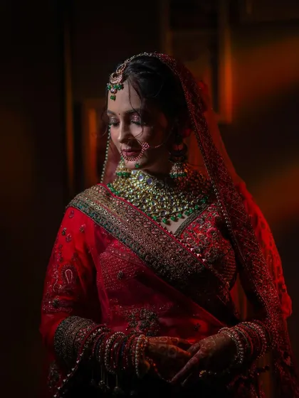 A classic bridal portrait showing the bride in her stunning red lehenga. The soft, dramatic lighting highlights her graceful pose and the richness of her wedding attire.
