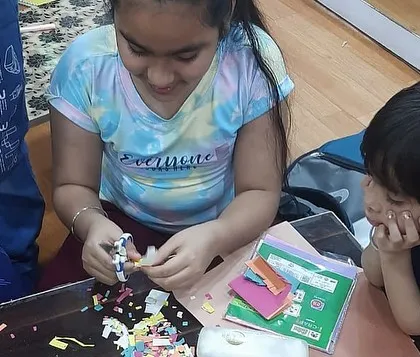 A candid shot of an older student helping a younger one during a craft session. Our camp environment fosters a sense of community and friendship.
