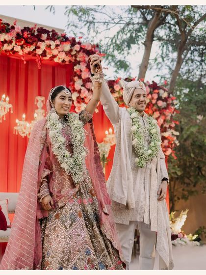 The couple celebrates their union on stage during the varmala ceremony, surrounded by a lush floral backdrop of red and white roses.
