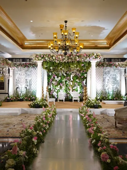 A beautiful and lush reception stage, with a mirrored aisle reflecting the floral-lined pathway and the grand backdrop of white florals and greenery.