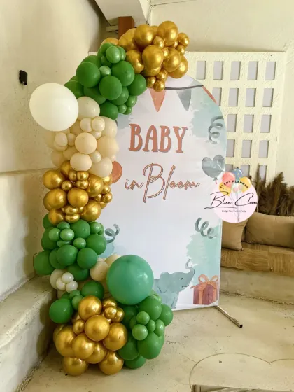 A 'Baby in Bloom' welcome sign with a green, gold, and white organic balloon garland.