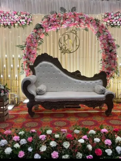 This stage setup features a circular floral arch with pink and white roses and a custom monogram of the couple's initials. The vintage-style chaise lounge adds a touch of royal elegance.