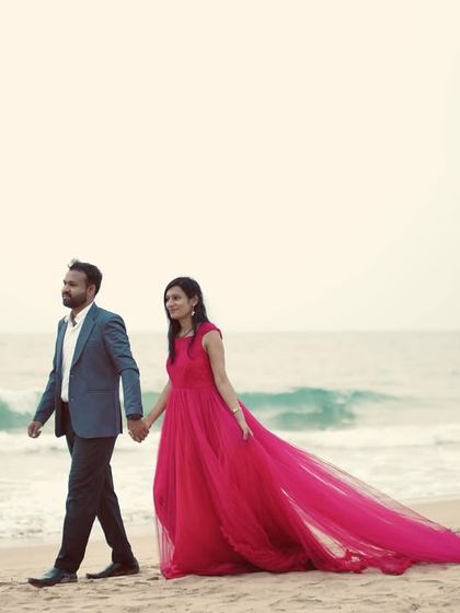 This wide shot captures the couple walking hand-in-hand on the beach, with the red trail gown adding a splash of color to the serene landscape.