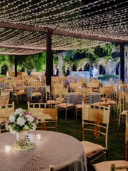 A closer view of the guest seating at the outdoor reception. The tables are dressed in striped linens with simple floral centerpieces, all under the magical glow of the fairy light canopy.