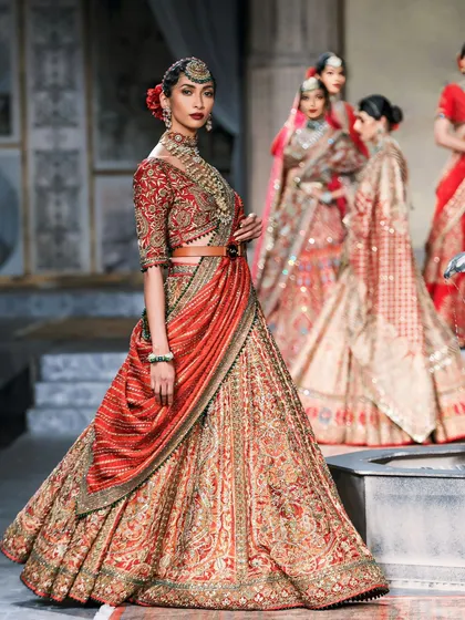 A model showcases a stunning red and gold bridal lehenga. The composition, with other models in the background, creates a rich, layered fashion photograph.
