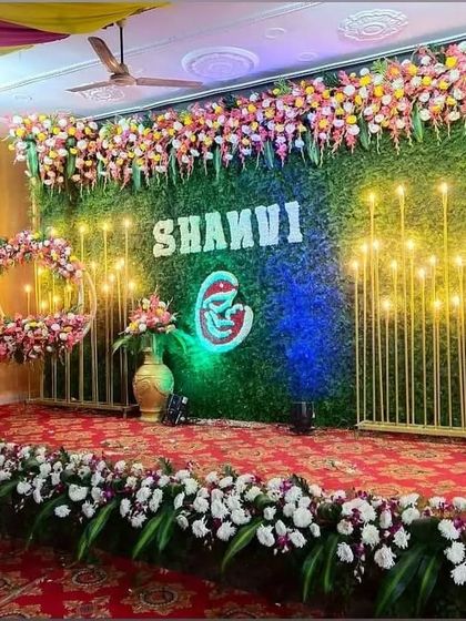 A beautiful stage for a naming ceremony with the baby's name "SHANVI" prominently displayed on a greenery wall, surrounded by flowers and lights.