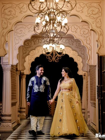 A royal pre-wedding shoot inside a heritage property. The couple walks down a corridor with a grand chandelier, their traditional outfits in yellow and blue creating a vibrant and regal scene.