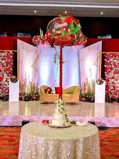 This modern reception stage combines marble-patterned panels with geometric gold frames and vibrant red floral arrangements. A custom hanging element with the couple's name adds a personalized and contemporary touch.