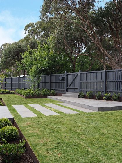 A view of the backyard showing the relationship between the lawn, the stepping stones, and the raised patio. The dark grey fence provides a neutral backdrop that makes the green foliage pop.