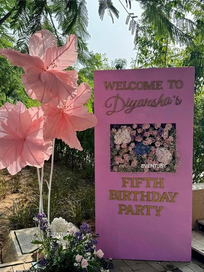 The welcome sign for the floral-themed fifth birthday party. The sign is decorated with large, handmade pink paper flowers, introducing the theme to guests as they arrive.