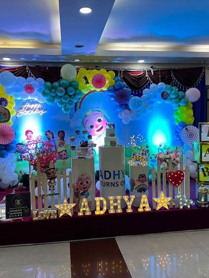 A colorful Cocomelon party stage with blue lighting effects. The setup includes a personalized backdrop, character cutouts, and a light-up marquee number '1'.