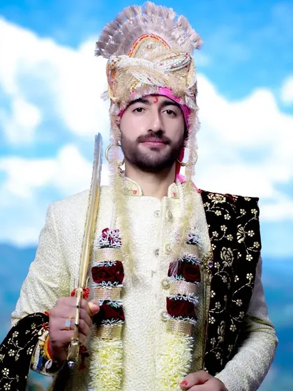A full-length portrait of the groom in his wedding sherwani, holding a ceremonial sword against a beautiful mountain sky.