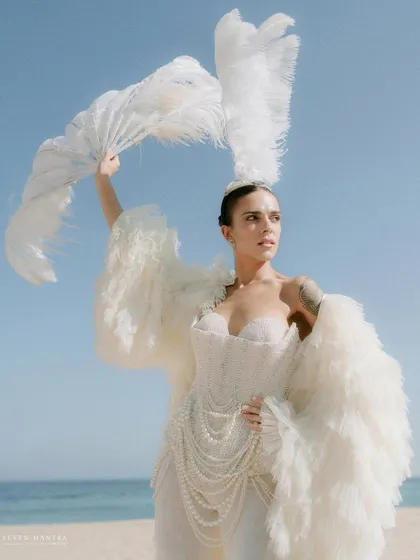 A goddess of the sea. This dramatic look, styled for a beach carnival, features a structured white corset, a flowing feather coat, and a dramatic feather fan, creating an unforgettable, high-fashion moment against the blue sky.