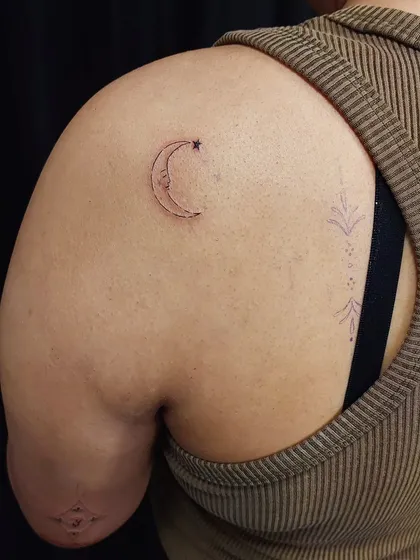 A whimsical crescent moon and star tattoo on the shoulder. The fine-line work gives it a delicate, hand-drawn feel.