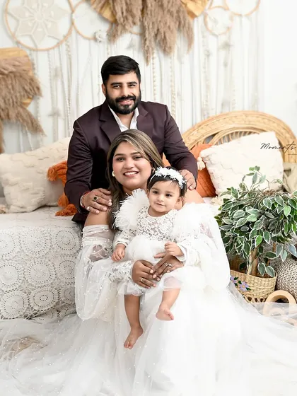 A serene family moment. The mother holds her daughter close while the father looks on, all dressed in white against a soft, bohemian backdrop.