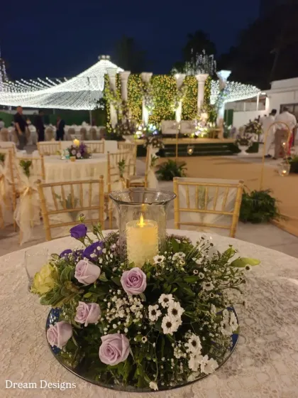 A close-up of a table centerpiece for the Parsi wedding. This arrangement features lavender roses and baby's breath, with a central candle in a glass hurricane lamp to provide a soft, romantic light.