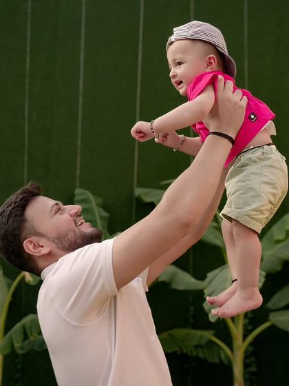 A father joyfully lifts his child into the air against a vibrant green backdrop.