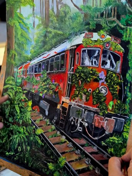 A close-up of the artist's hands adding fine details to the train and surrounding greenery in her gouache artwork.