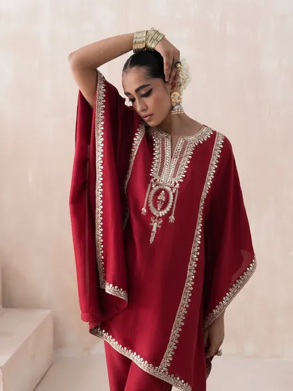 A close-up shot for Saisha's "Firdaus" collection, featuring a red kaftan-style kurta with intricate gold embroidery. The styling includes traditional jewelry and a floral hair accessory to complete the elegant look.