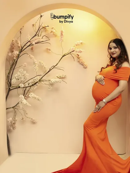 A vibrant and modern studio portrait. The mom-to-be looks stunning in a bright orange form-fitting gown, posed against our minimalist floral arch set.