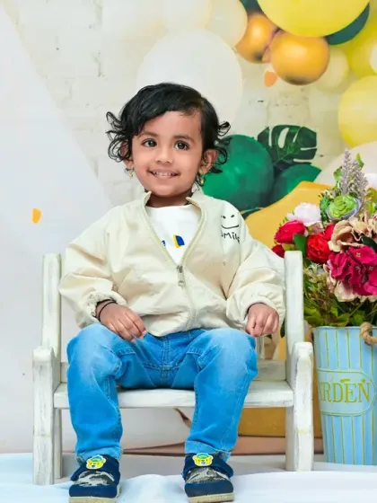 This happy toddler is celebrating his second birthday in style. Sitting proudly in a little white chair, he smiles for the camera, with "TWO" blocks and a festive balloon arch setting the scene.