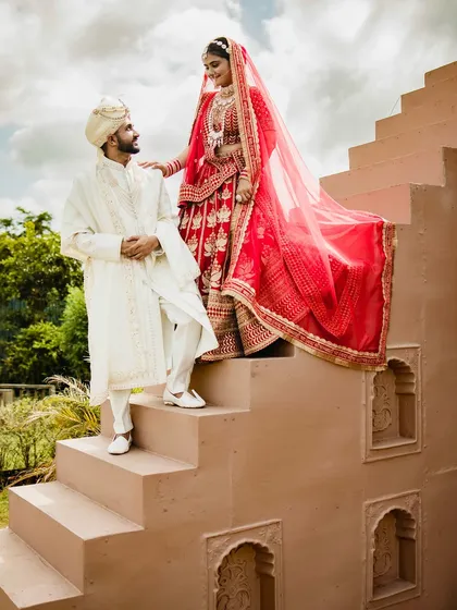 A creative pose on the steps of the venue. The bride stands above the groom, her red lehenga flowing down, creating a visually interesting and dynamic portrait.
